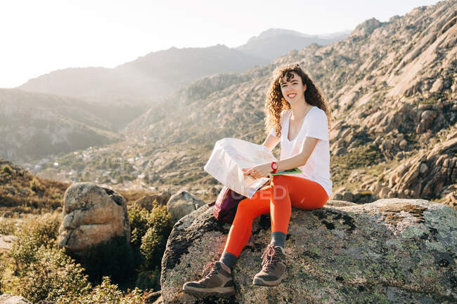 Joven excursionista femenina positiva con el pelo rizado sentado en la roca de piedra y sonriendo mientras lee un mapa para la orientación en el valle montañoso en el día soleado - foto de stock