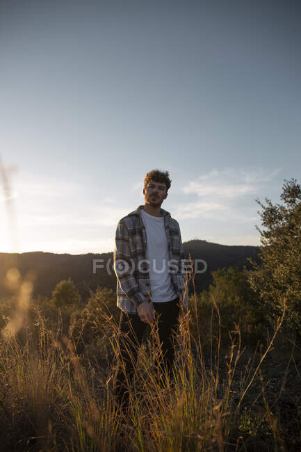 Небритый мужчина в клетчатой рубашке стоит среди выцветшей травы под мягким солнечным светом вечером — стоковое фото