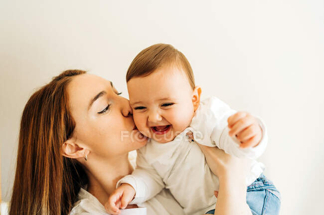 Молодая мать в повседневной одежде обнимает и целует маленького ребенка в светлой комнате — стоковое фото