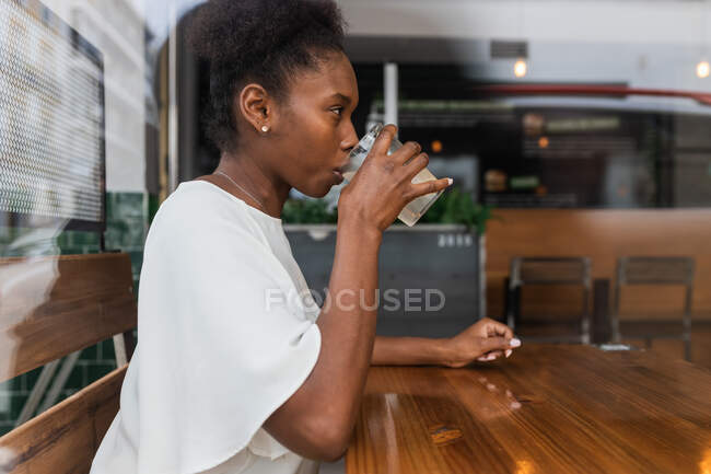 Vista laterale di donna afroamericana calma in abito casual bere acqua fresca fredda dalla bottiglia in vetro con ghiaccio e limone mentre seduto al tavolo alto in caffetteria — Foto stock