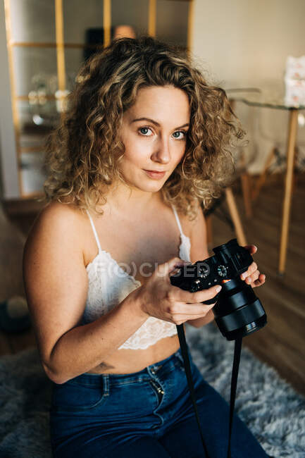 Молодая сконцентрированная женщина-фотограф с вьющимися светлыми волосами в кружевном бюстгальтере и джинсах сидит на мягком ковре и фотографирует дома на камеру — стоковое фото