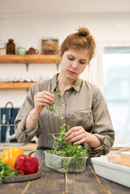 Молода жінка зі свіжими зеленими травами в пластиковій коробці за столом з перцем готує вдома — стокове фото