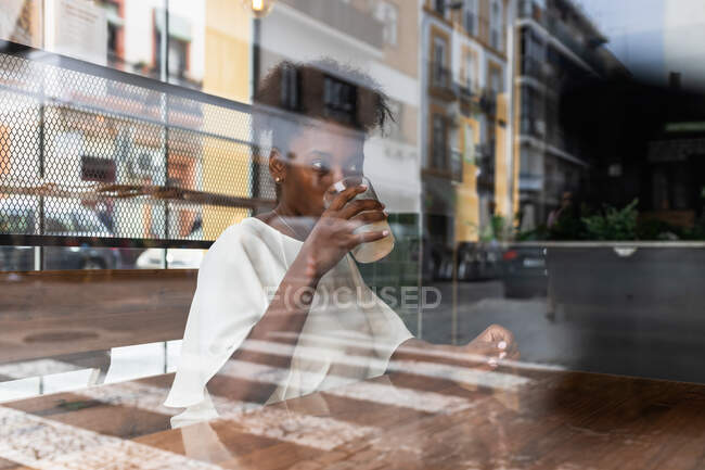 Through glass crop calm African American female in casual outfit drinking cold fresh water from bottle into glass with ice and lemon while sitting at high table in cafeteria — Stock Photo