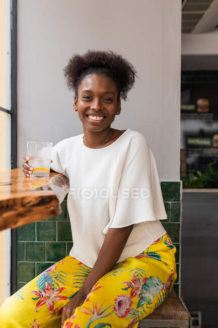 Side view of happy African American female in casual outfit drinking cold fresh water from bottle into glass with ice and lemon while sitting at high table in cafeteria while looking at camera — Stock Photo