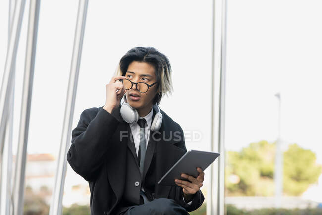 Joven empresario masculino étnico en auriculares modernos y gafas con tableta mirando hacia otro lado en la ciudad - foto de stock