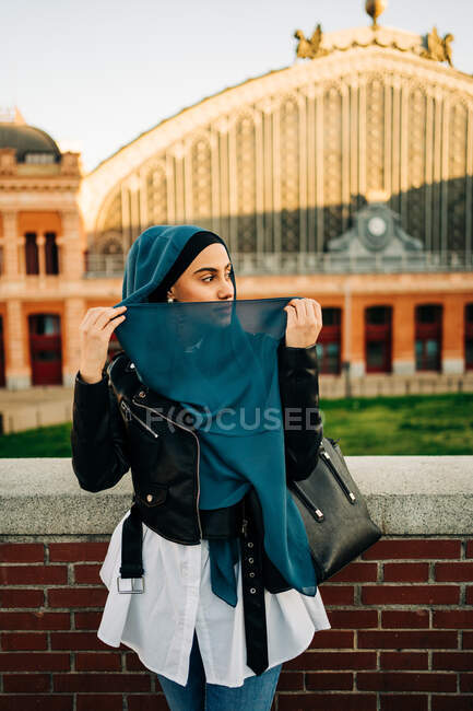 Contenida mujer étnica en hijab tradicional y ropa de moda de pie en la calle de la ciudad y mirando hacia otro lado - foto de stock