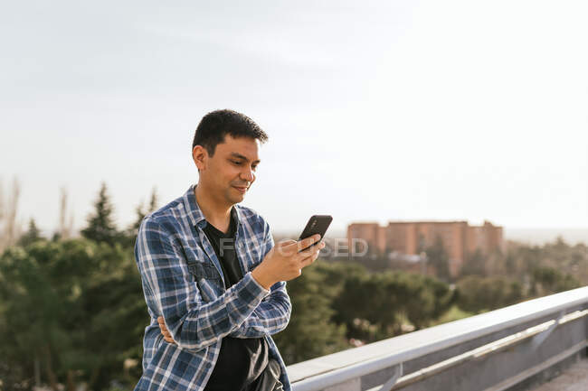Konzentrierter Mann mit Brille und kariertem Hemd, der an einem sonnigen Sommertag auf dem Dach eines Vorortes steht und im modernen Mobiltelefon surft — Stockfoto