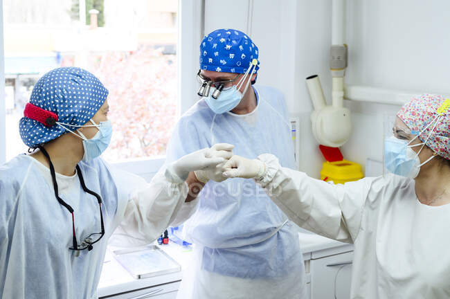 Cirujano masculino con compañeras de trabajo en uniforme saludándose con los cinco altos en el trabajo en el hospital - foto de stock