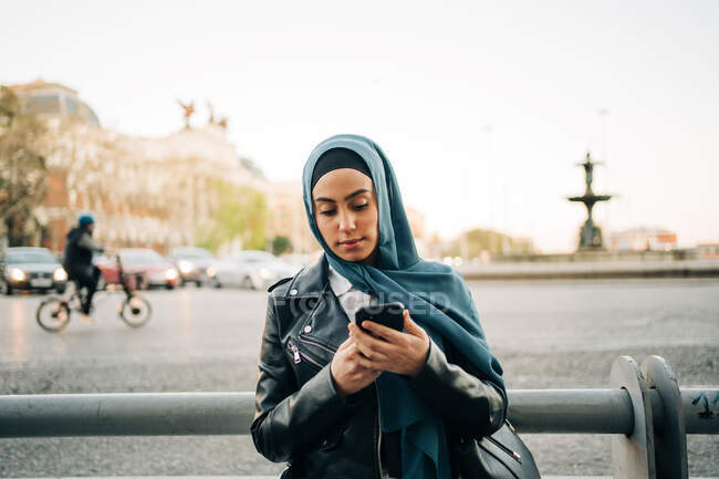 Mujer musulmana en pañuelo de cabeza de pie en la calle de la ciudad y el teléfono móvil de navegación - foto de stock