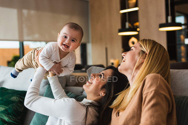 Alegre pareja de mujeres homosexuales jugando con lindo niño mientras se divierten y disfrutan de fin de semana juntos en casa - foto de stock