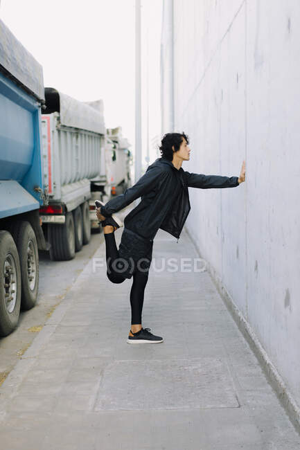 Vista lateral macho de corpo inteiro em roupas esportivas pretas totais inclinadas com a mão na parede enquanto se alonga no distrito industrial da cidade — Fotografia de Stock