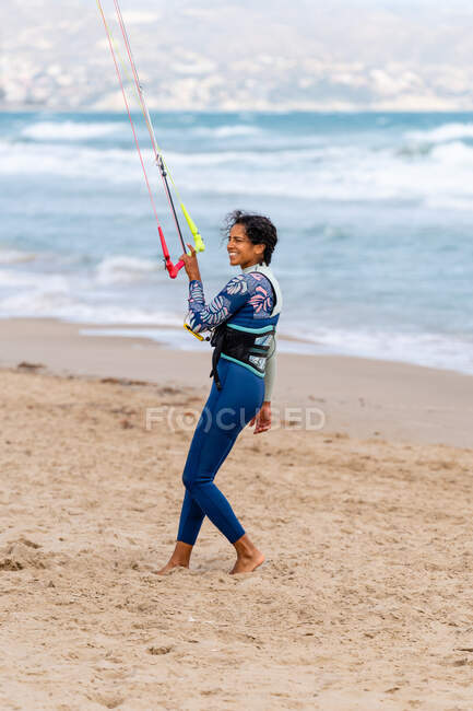 Vista lateral de kiter feminino alegre em wetsuit segurando barra de controle enquanto olha para longe na costa do oceano arenoso — Fotografia de Stock