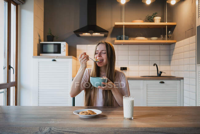 Jovem fêmea com colher e tigela desfrutando saborosos anéis de milho na cozinha — Fotografia de Stock