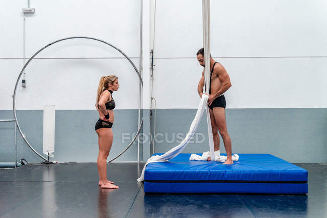 Acróbatas descalzos de cuerpo completo en ropa interior deportiva preparándose para entrenar con seda aérea en un moderno estudio de gimnasia - foto de stock