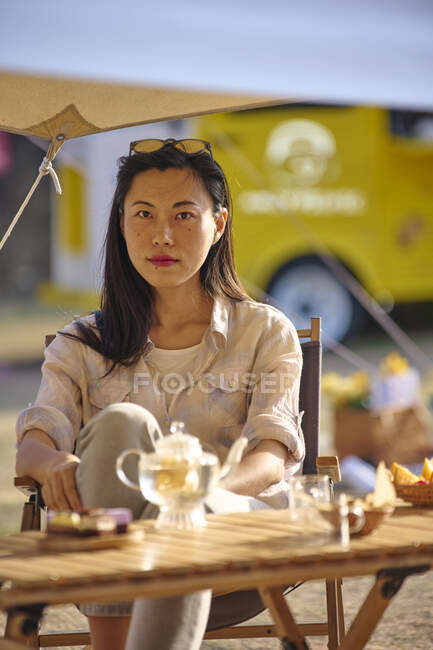 Linda étnica asiática fêmea sentada à mesa enquanto tendo um tempo relaxante na área de acampamento durante as férias olhando para a câmera — Fotografia de Stock
