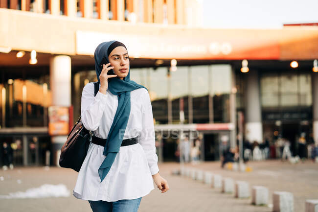 Donna etnica in hijab in piedi vicino alla stazione ferroviaria e parlando cellulare mentre distoglie lo sguardo — Foto stock