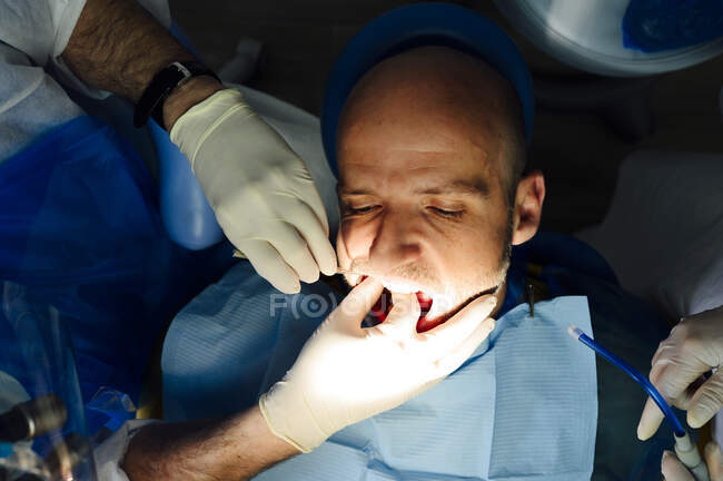 Cosecha ortodoncista anónimo en guantes de látex con compañero de trabajo comprobando los dientes de hombre maduro mirando hacia adelante en el hospital - foto de stock