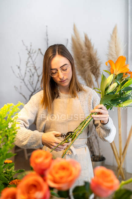 Focused young ethnic female florist in casual clothes and apron cutting stems of fresh bright flowers with pruning snips in floral shop — Stock Photo