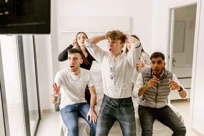 Group of expressive young multiethnic friends in casual clothes sitting on comfortable bed and watching sport match together on TV during weekend at home — Foto stock