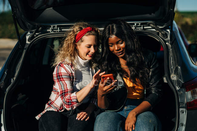 Щасливі багаторасові друзі, що сидять у багажнику автомобіля та переглядають соціальні мережі на мобільному телефоні разом — стокове фото