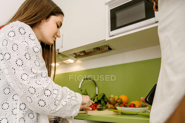 Low angle of crop ethnic female in stylish outfit cutting fresh strawberry on chopping board while cooking in kitchen with cropped unrecognizable boyfriend — Fotografia de Stock