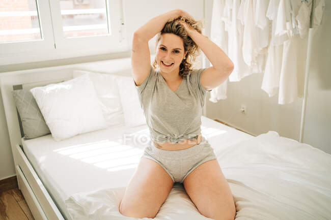 Sensual joven hembra en la parte superior y bragas tocando el pelo rizado y sonriendo mientras se sienta en la cama cómoda durante el fin de semana en casa - foto de stock