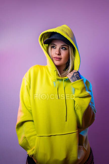 Unemotional female in street style hoodie and cap looking away on purple background in studio with neon illumination — Stock Photo