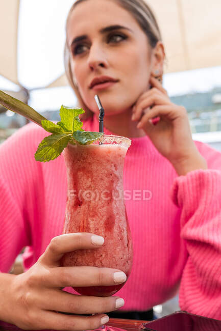 Mulher sonhadora da colheita em suéter rosa sentado à mesa com delicioso coquetel de frutas frias no bar ao ar livre e olhando para longe — Fotografia de Stock