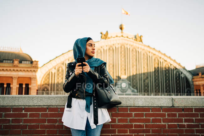Mulher muçulmana em lenço de cabeça tradicional em pé na rua da cidade e telefone de navegação enquanto olha para longe — Fotografia de Stock