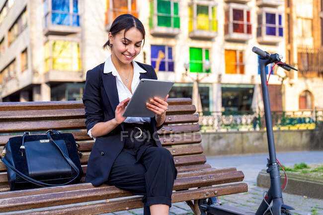 Conteúdo jovem empresária étnica sentada no banco enquanto navega no tablet perto de scooter elétrico e edifícios da cidade — Fotografia de Stock