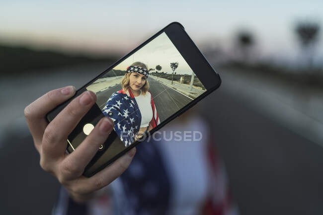 Патриотичная американка, завернутая в национальный флаг США, делает селфи на мобильном телефоне, стоя на дороге — стоковое фото