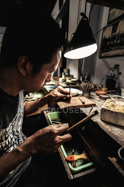 Side view of ethnic craftsman looking close at metal piece and punching metal stamp with small hammer — Stock Photo
