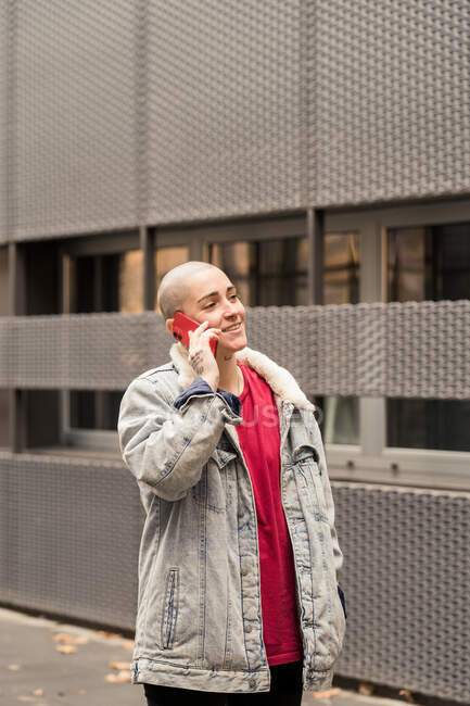Трансгендер в повседневной одежде разговаривает по мобильному телефону, глядя в сторону городского здания при дневном свете — стоковое фото