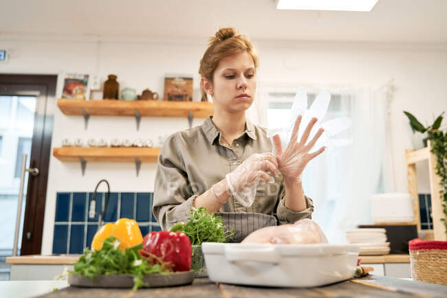 Female putting on transparent gloves and looking down against fresh peppers with herbs and raw chicken in baking pan at home — Stock Photo