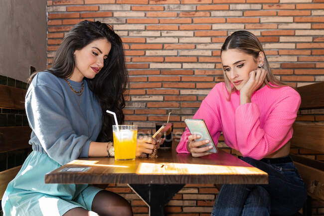 Jovens amigas elegantes navegando telefones celulares enquanto se sentam à mesa com bebidas na cafetaria — Fotografia de Stock