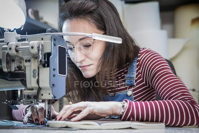 Artesana enfocada usando máquina de coser mientras crea tapicería para asiento de moto en taller - foto de stock