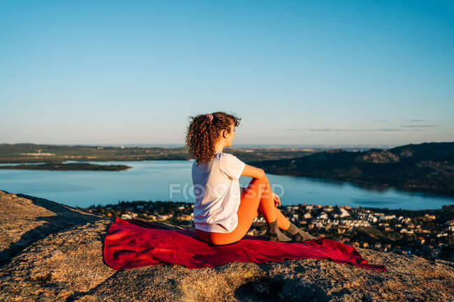 Vue latérale d'une jeune voyageuse enchantée aux longs cheveux foncés en tenue décontractée assise sur une falaise rocheuse et admirant la mer contre un ciel sans nuages au coucher du soleil — Photo de stock