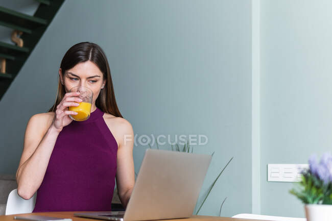 Attractive smiling female in casual top using modern netbook while sitting at table with glass of fresh orange juice — Stock Photo