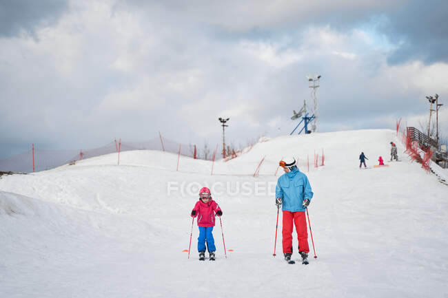 Повне тіло безликого батька в теплому спортивному одязі та шоломі навчає маленьку дитину кататися на лижах уздовж сніжного схилу пагорба в зимовому гірськолижному курорті — стокове фото