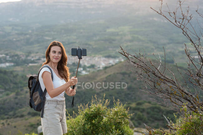 Vista lateral da fêmea viajante com mochila em pé na colina e tomando auto-tiro no smartphone no fundo da gama de montanhas no verão — Fotografia de Stock