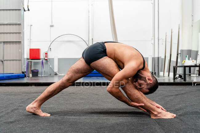 Полное тело подходит гибкому мужчине в спортивных шортах, делающему треугольную позу и смотрящему в камеру, практикуя йогу в современном фитнес-центре — стоковое фото