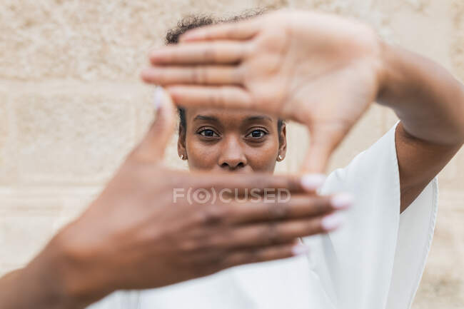 Happy young African American female in white blouse showing on camera framing sign with hands and looking at camera while standing against uneven wall — Stock Photo