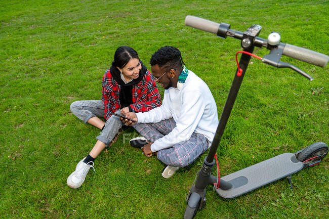 From above of cheerful multiracial couple in trendy wear with cellphone speaking on meadow with electric scooter in daytime — Stock Photo