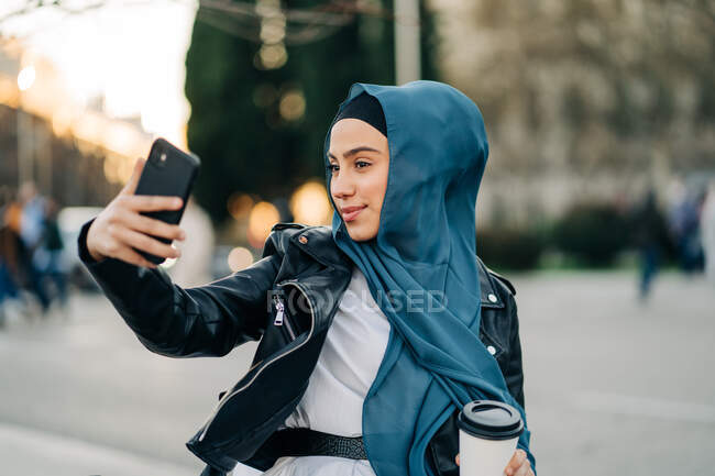Femme ethnique joyeuse portant le foulard traditionnel debout dans la rue avec boisson à emporter et prenant l'autoportrait sur smartphone — Photo de stock