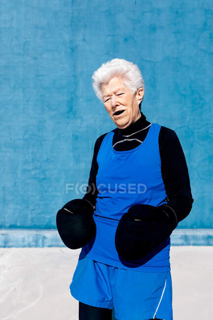 Cheerful elderly female in activewear in boxing gloves against blue wall and looking at camera — Stock Photo