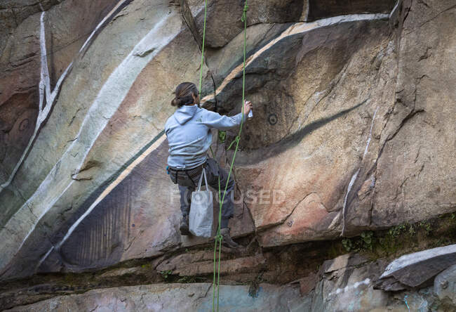 Back view full body of painter with spray paint making graffiti hanging on rope on steep rocky slope — Stock Photo