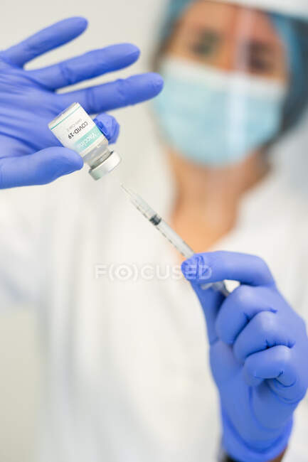 Médecin en gants de latex et bouclier facial remplissant la seringue du flacon avec le vaccin se préparant à vacciner le patient en clinique pendant l'épidémie de coronavirus — Photo de stock