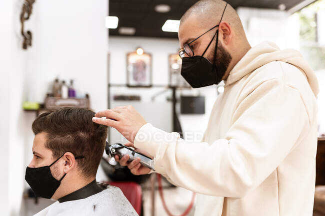 Peluquero masculino en anteojos haciendo corte de pelo al cliente adulto en peluquería durante la pandemia de COVID 19 - foto de stock