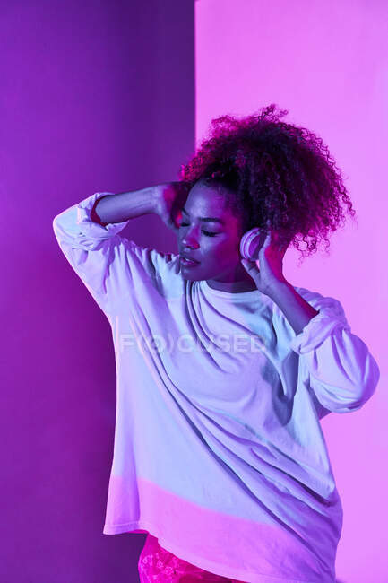 Jeune femme afro-américaine réfléchie en vêtements blancs portant un casque sur le cou tout en se tenant les yeux fermés dans un studio sombre aux néons — Photo de stock