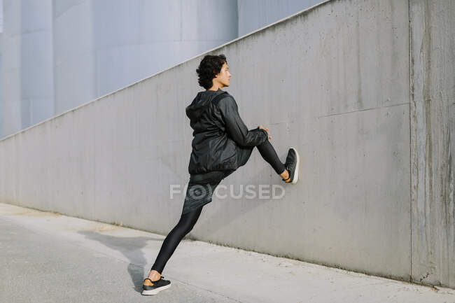 Side view full body male in total black sportive clothes leaning with leg on bock while stretching in industrial city district — Stock Photo